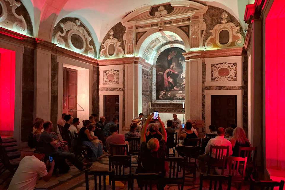 A small musical performance in a historic hall with audience seated, ornate walls, and a large painting in the background.