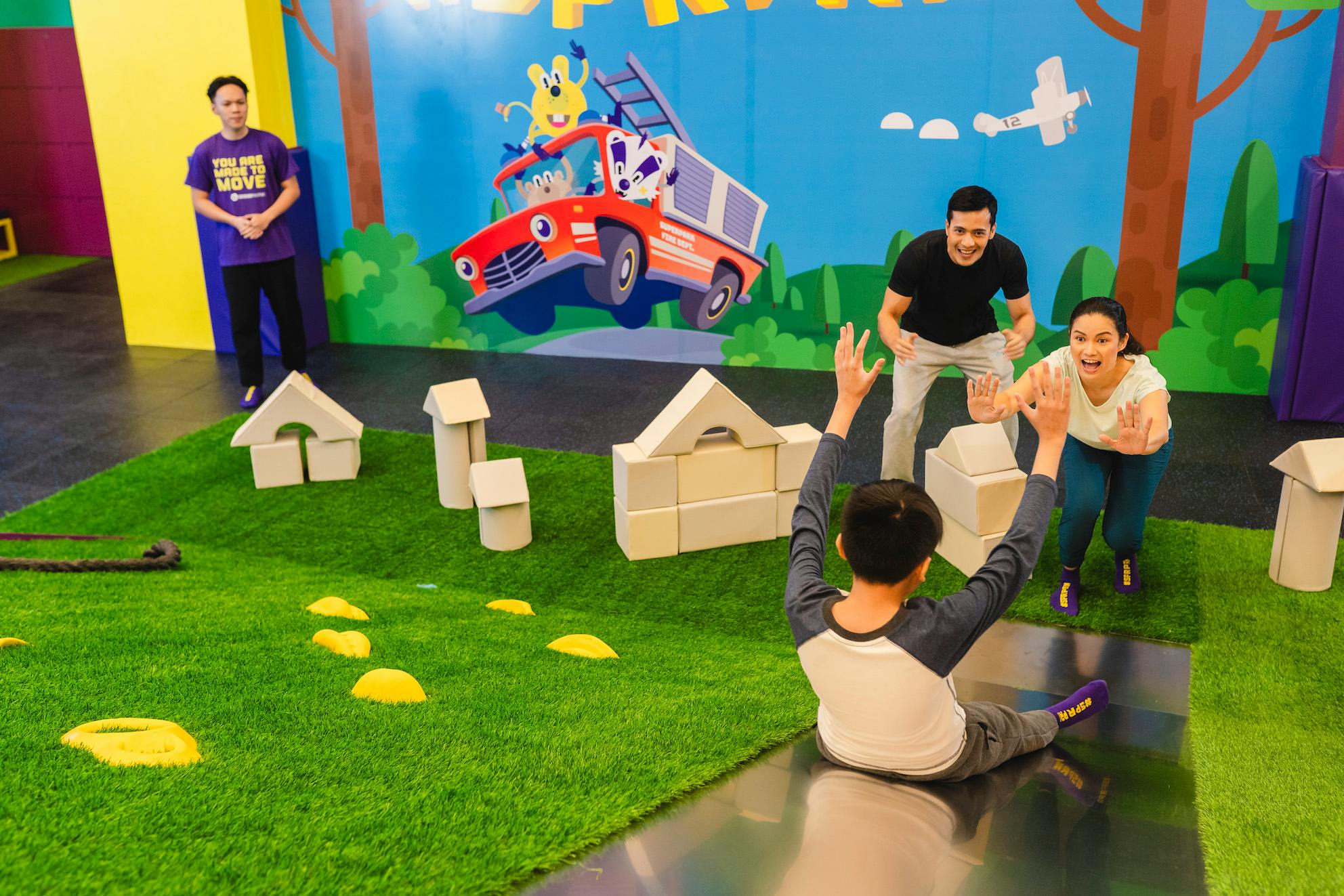 Children and adults playing with foam blocks on artificial grass in a colorful indoor play area with a mural on the wall.