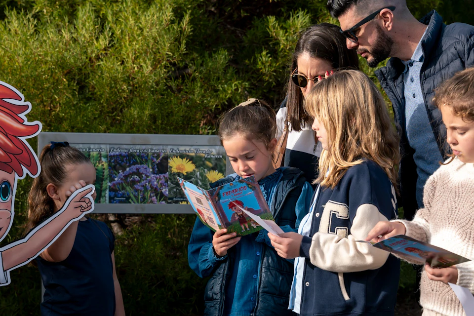 A group of children and an adult stand outdoors reading books near a colorful sign. Trees are visible in the background.
