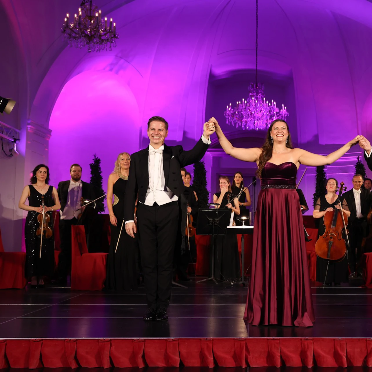 Two performers in formal attire hold hands on a stage, with a symphony orchestra and purple-lit background.