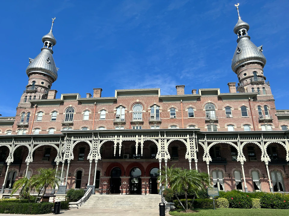 Historic building with ornate arches, two silver-domed towers, and a lush garden under a bright blue sky.