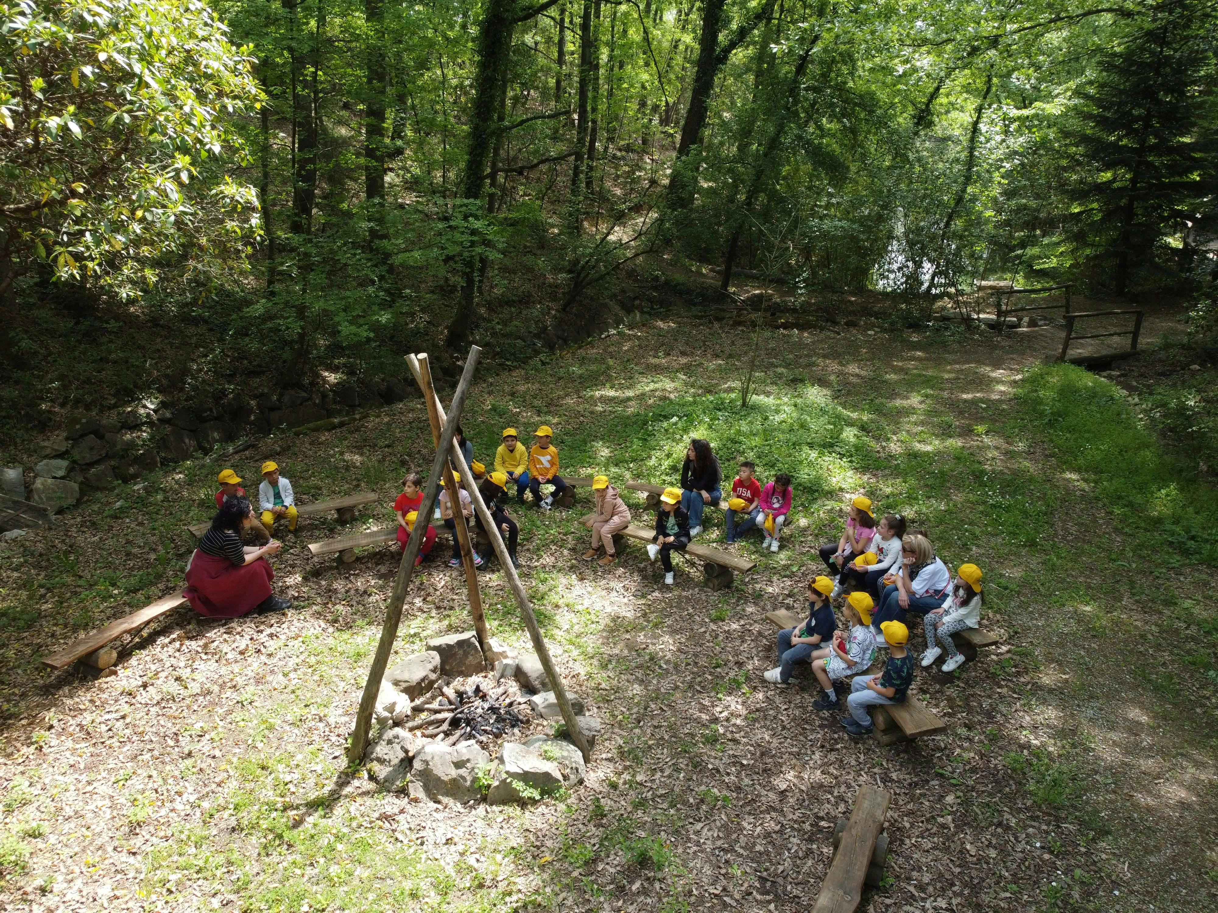 孩子们坐在森林空地上的长凳上，在临时搭建的篝火旁仔细聆听两名成年人的声音。