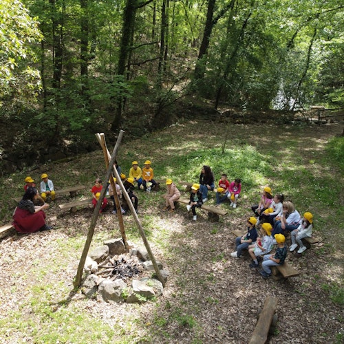 I bambini siedono su panchine in una radura della foresta, ascoltando attentamente due adulti vicino a un falò improvvisato.
