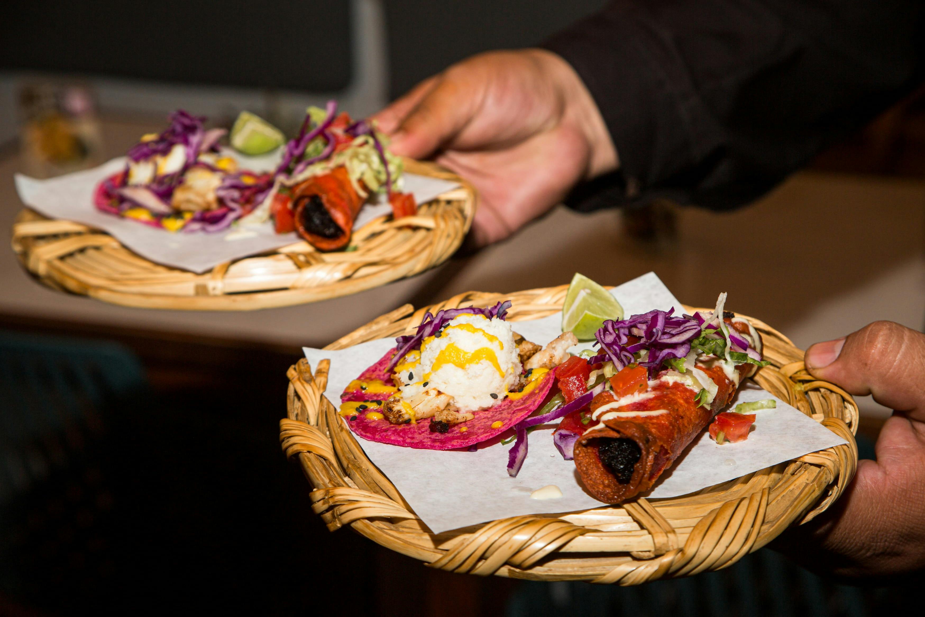 Una persona tiene in mano due cestini intrecciati contenenti tacos al piatto con guarnizioni e condimenti colorati, serviti su carta pergamena.