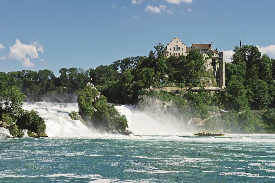 A scenic waterfall flows with water surrounded by greenery and buildings on a sunny day under a blue sky.