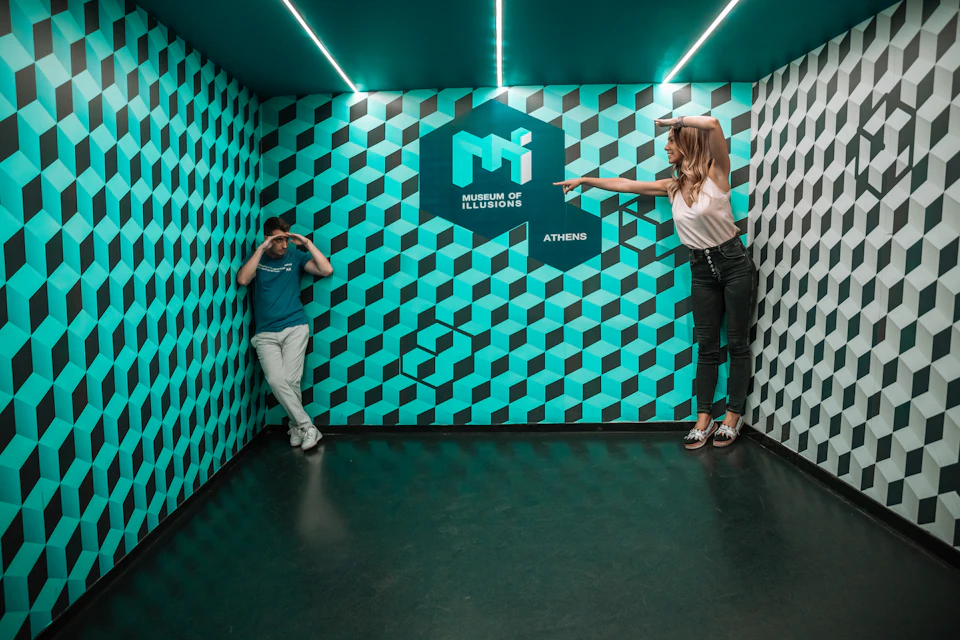 Two people in a room with a geometric optical illusion wall at the Museum of Illusions in Athens. The woman points at the man.