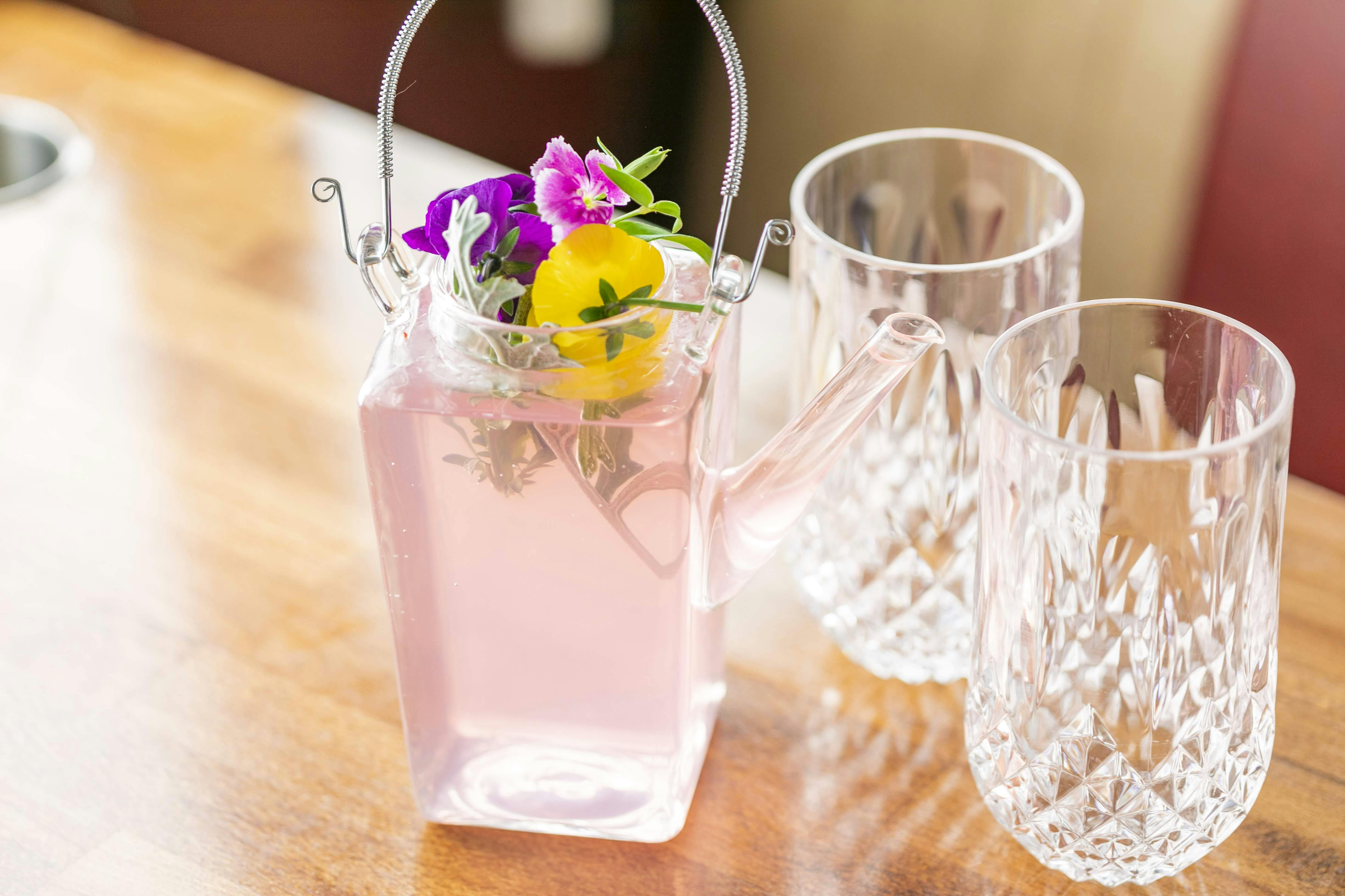 Une théière en verre avec un liquide rose et des fleurs colorées, à côté de deux verres en cristal sur une table en bois.
