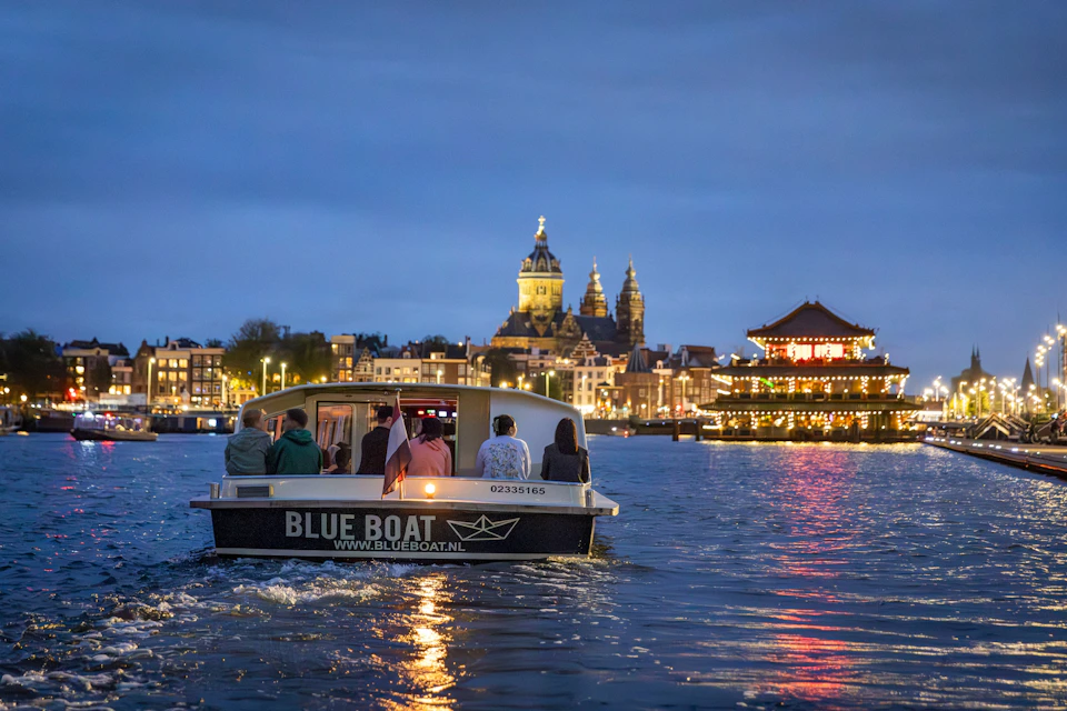 A boat labeled 'Blue Boat' with passengers on a waterway, a lit cityscape with a church and buildings visible at dusk.