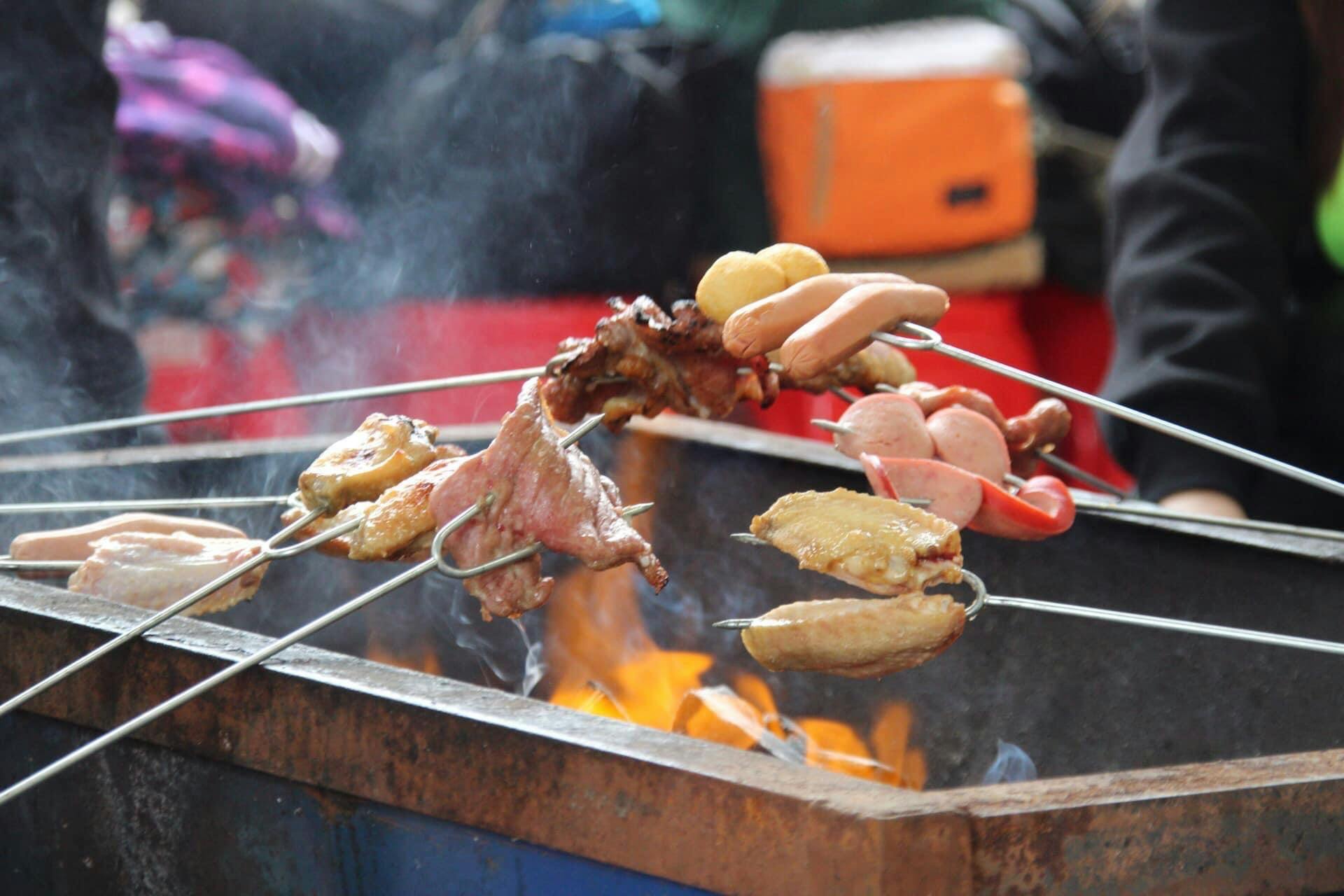 Skewers with various meats and vegetables cooking over an open flame on a metal grill, with blurred background.