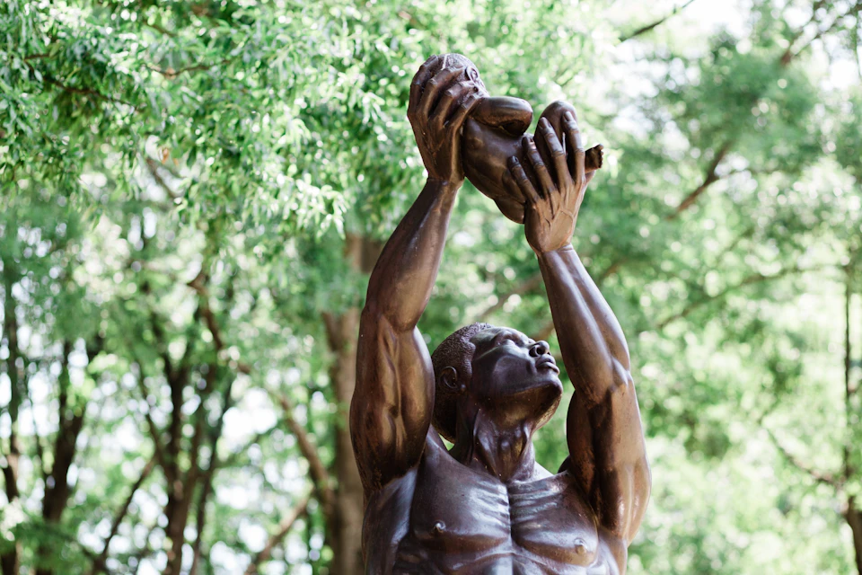 Bronze statue of a man holding a child above his head, with trees in the background.