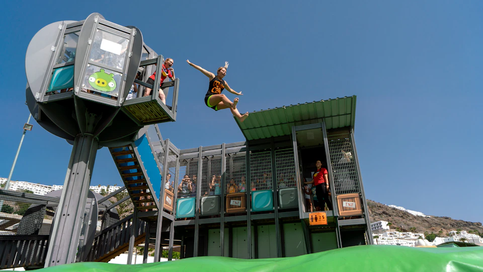 Person in mid-air jumping from a platform at an adventure park, with onlookers and a clear blue sky in the background.