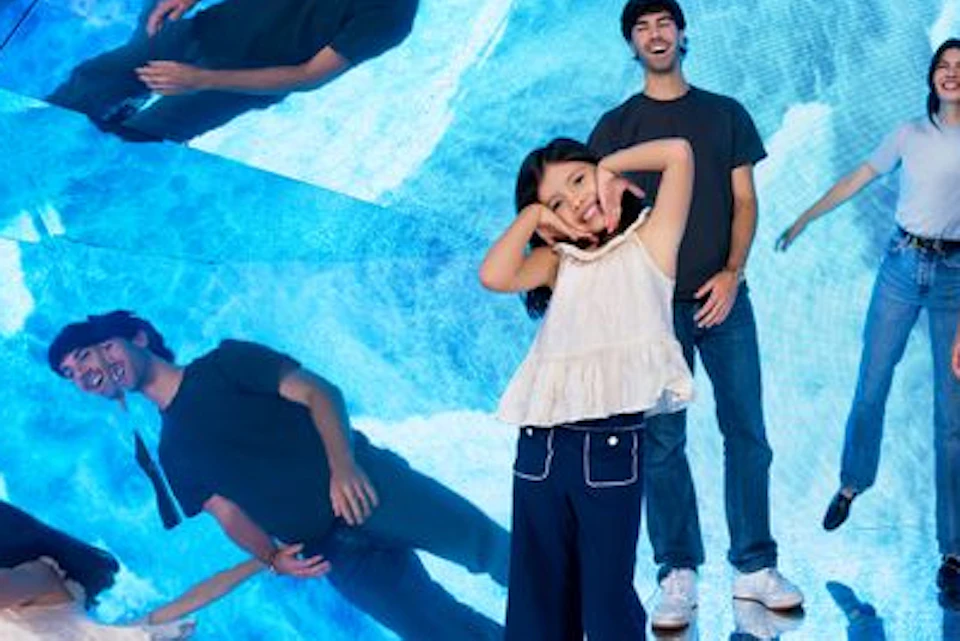 A group of people posing in a room with walls covered in blue mirror-like reflections, creating a kaleidoscope effect.