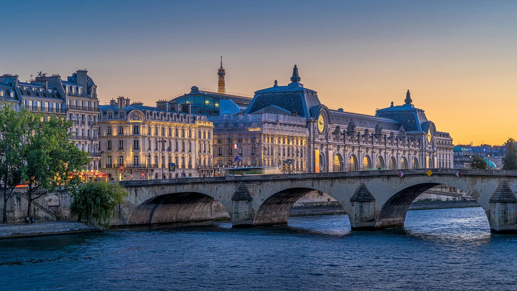 Parisian cityscape at sunset with historic buildings, a bridge over the Seine, and the Eiffel Tower in the background.