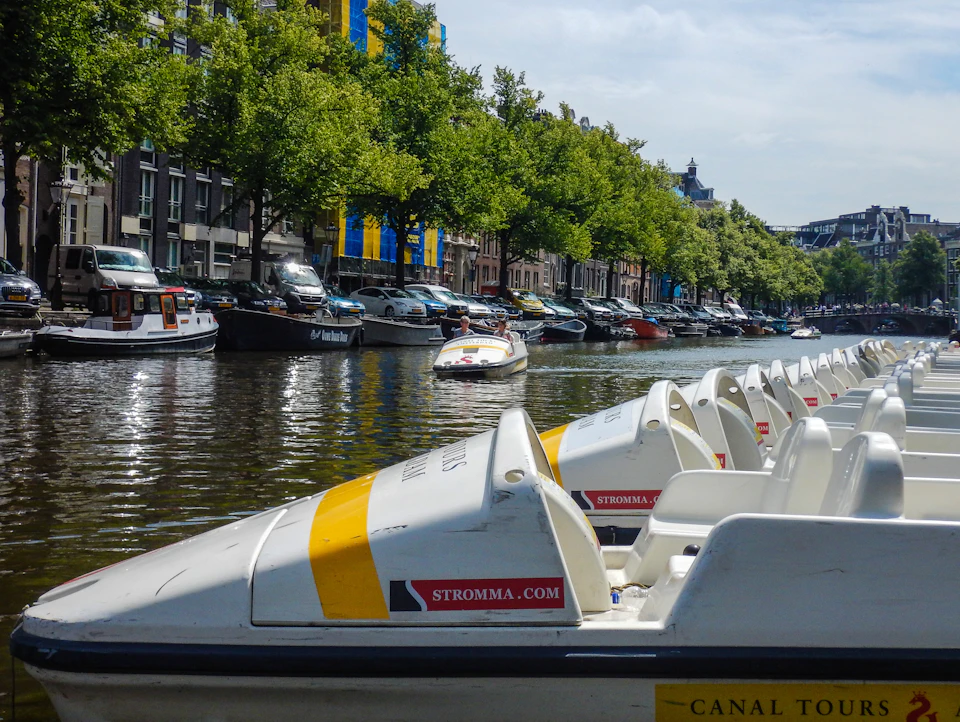 Pedal boats lined up on a canal with larger boats and trees in the background, and a boat navigating the water.