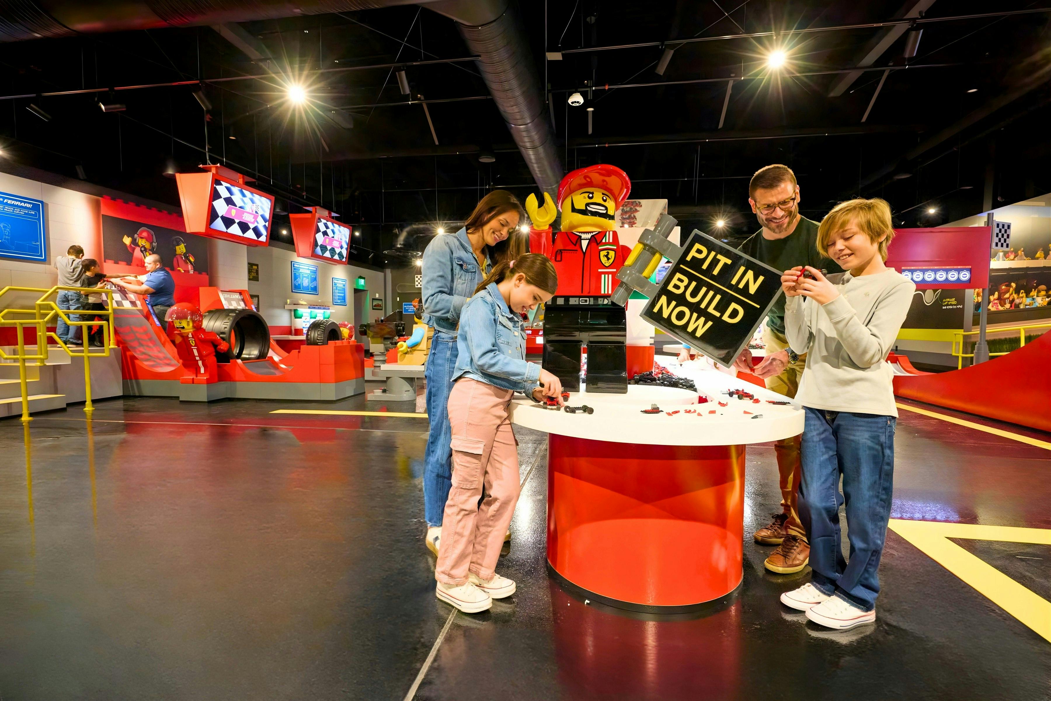 Una família interactua amb els maons de Lego en una taula d'un centre d'activitats interior, amb una gran figura de Lego al fons.