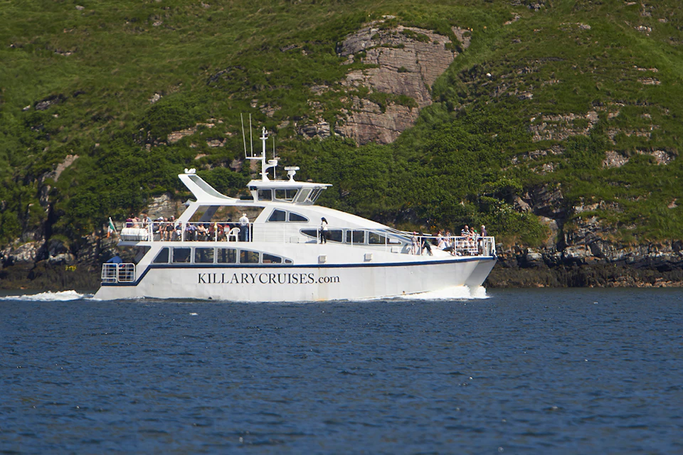 A white Killary Cruises boat with passengers on deck sails near a rocky, green hillside.