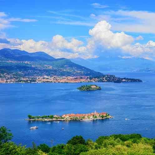 Vue aérienne d'un lac avec de petites îles, des bateaux et une ville côtière, sur fond de collines et de ciel partiellement nuageux.