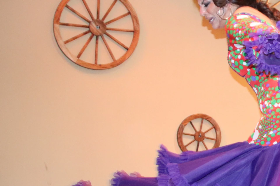 A woman in a colorful dress performs a dance on stage, with a man applauding in the background. Decor includes wheel decor on the walls.