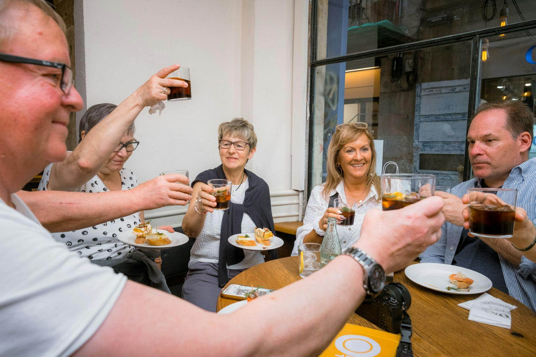 Um grupo de pessoas sentadas à mesa, a sorrir e a brindar com bebidas, com pratos de comida à sua frente.