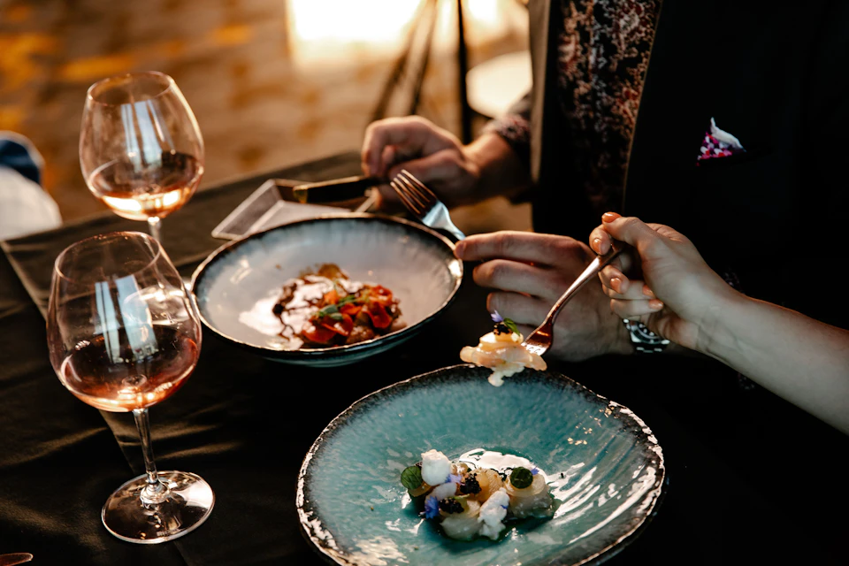 Two people dining with gourmet dishes on plates and glasses of wine, one holding a fork with food over a blue plate.