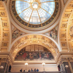 Stora salen med ett välvt glastak, väggmålningar, utsmyckade valv och gyllene detaljer. Människor står på en balkong nedanför.