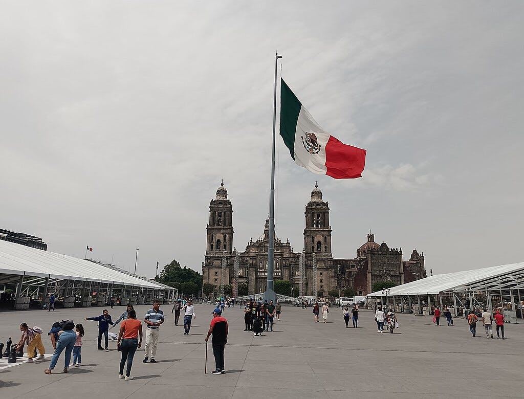 People walking in a large plaza with a tall Mexican flag and a historic cathedral in the background.