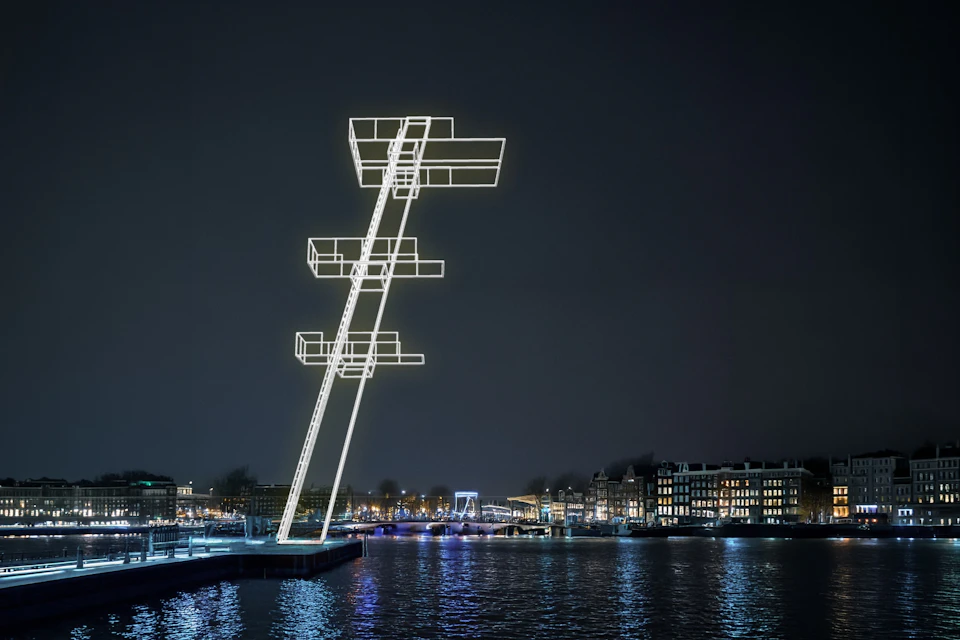 A glowing art installation resembling stacked platforms over water at night, with city buildings in the background.