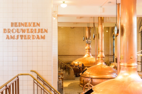 왼쪽 벽에 구리 양조 탱크, 흰색 타일 벽, "Heineken Brouwerijen Amsterdam" 텍스트가 있는 양조장 내부.