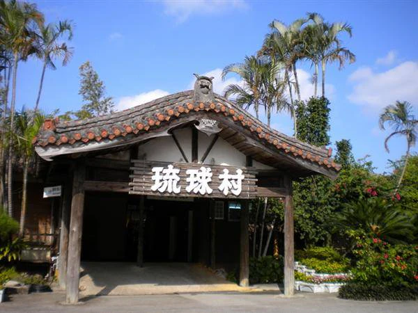 Traditional wooden gate with Japanese characters under a tiled roof, surrounded by trees on a sunny day.