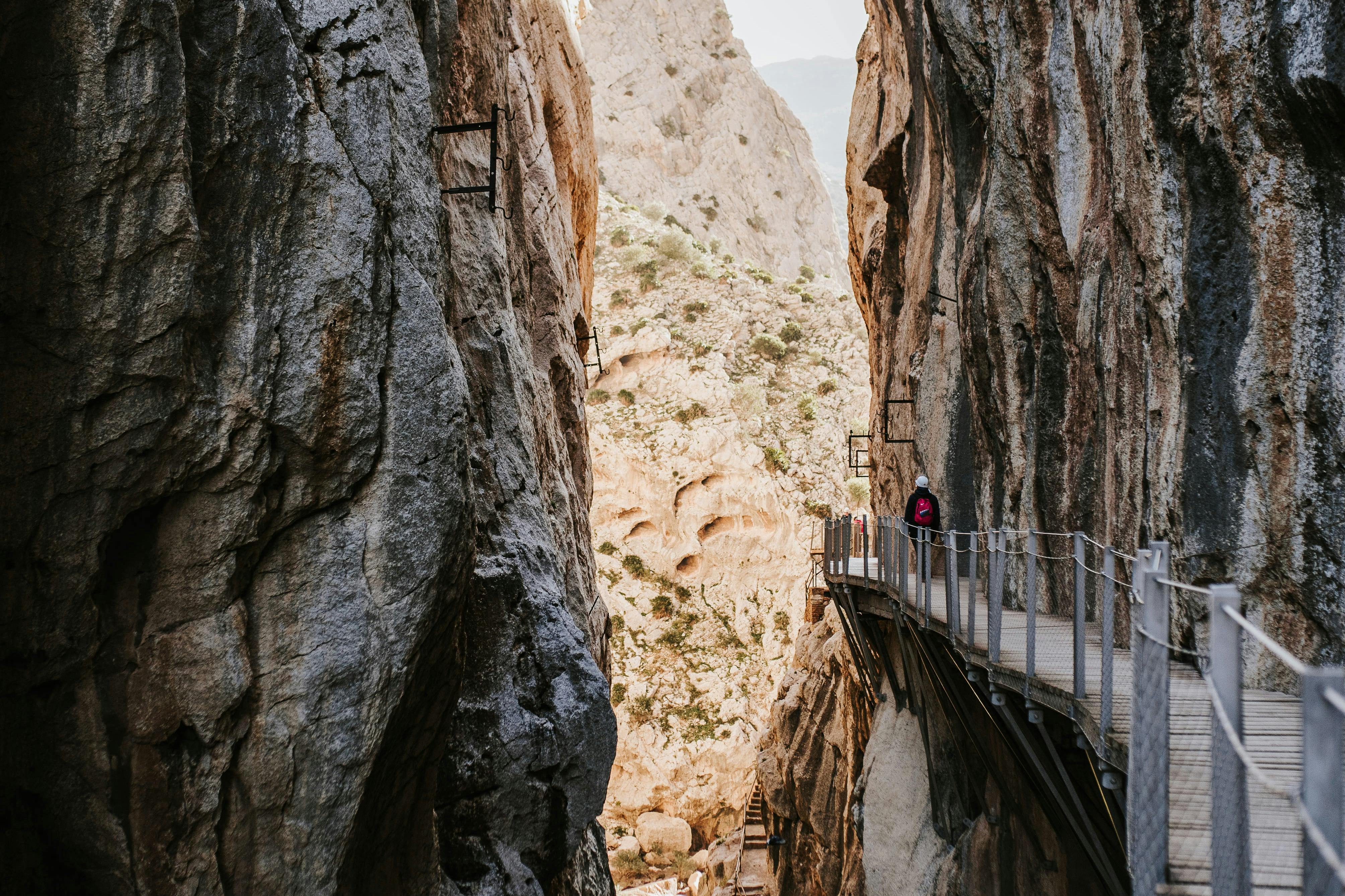 一条狭窄的小径，两座高耸的岩石悬崖之间悬挂着栏杆，一个人走在小径上。