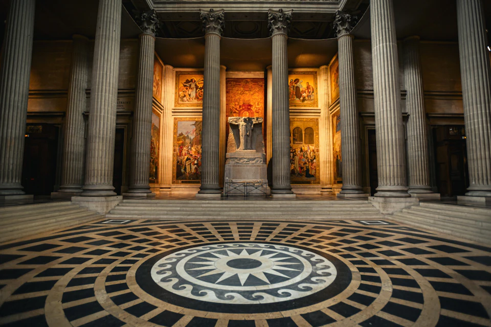 A grand hall with ornate columns, intricate floor mosaic, and illuminated wall murals surrounding a central statue.
