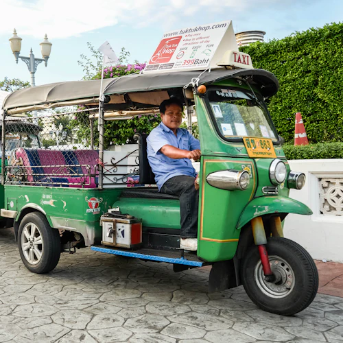 Ein Mann fährt ein grünes Tuk-Tuk mit Passagierwagen, das vor einem traditionellen thailändischen Gebäude geparkt ist.