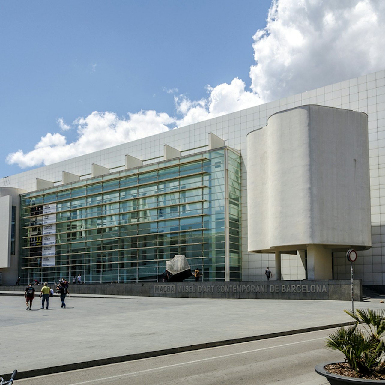 Modern vit byggnad med glasfasad, uteservering med människor och små palmplantor i förgrunden. Klarblå himmel.