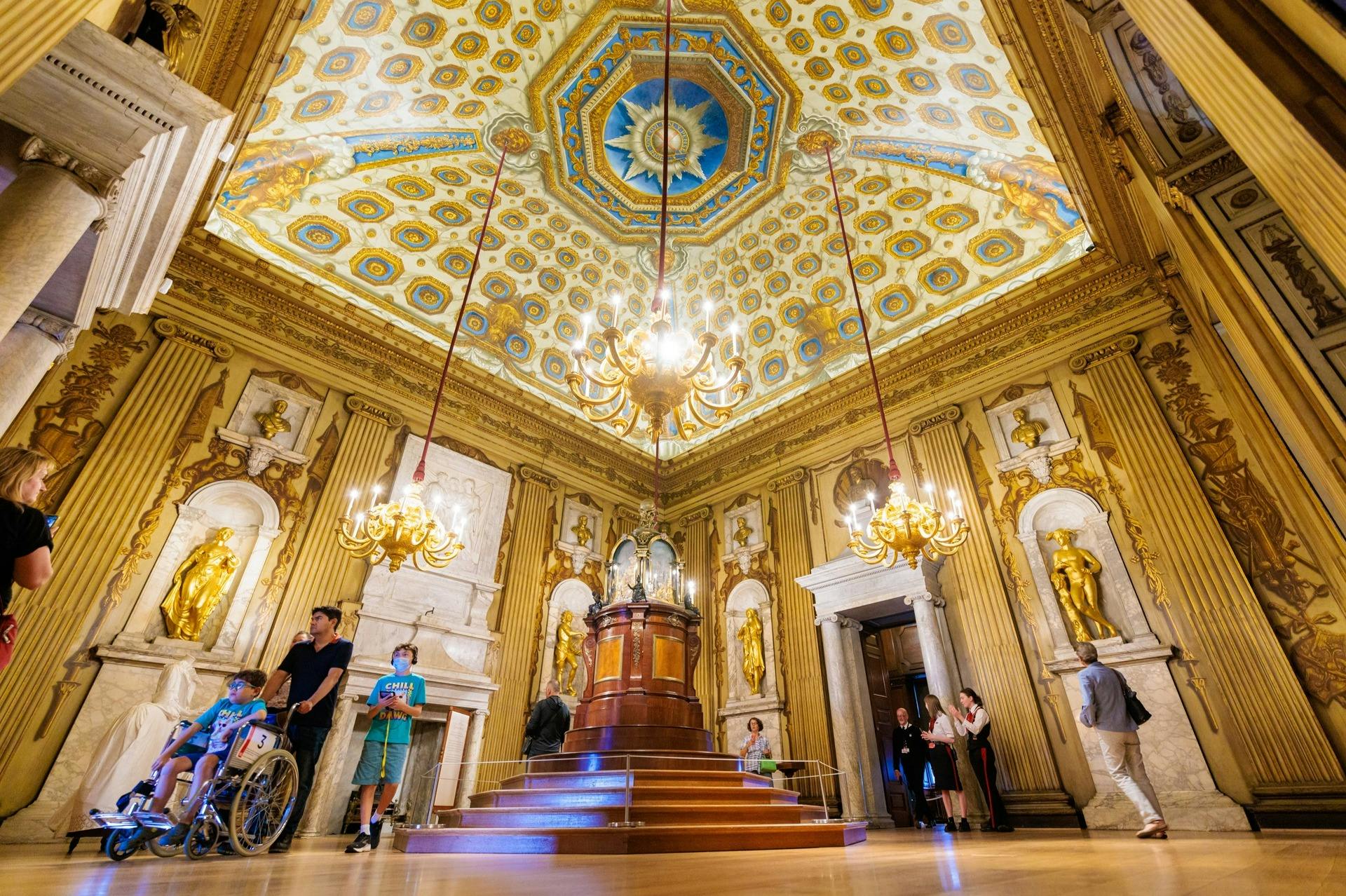 Visitors explore an ornate room with a decorated ceiling, chandeliers, statues, and intricate woodwork.