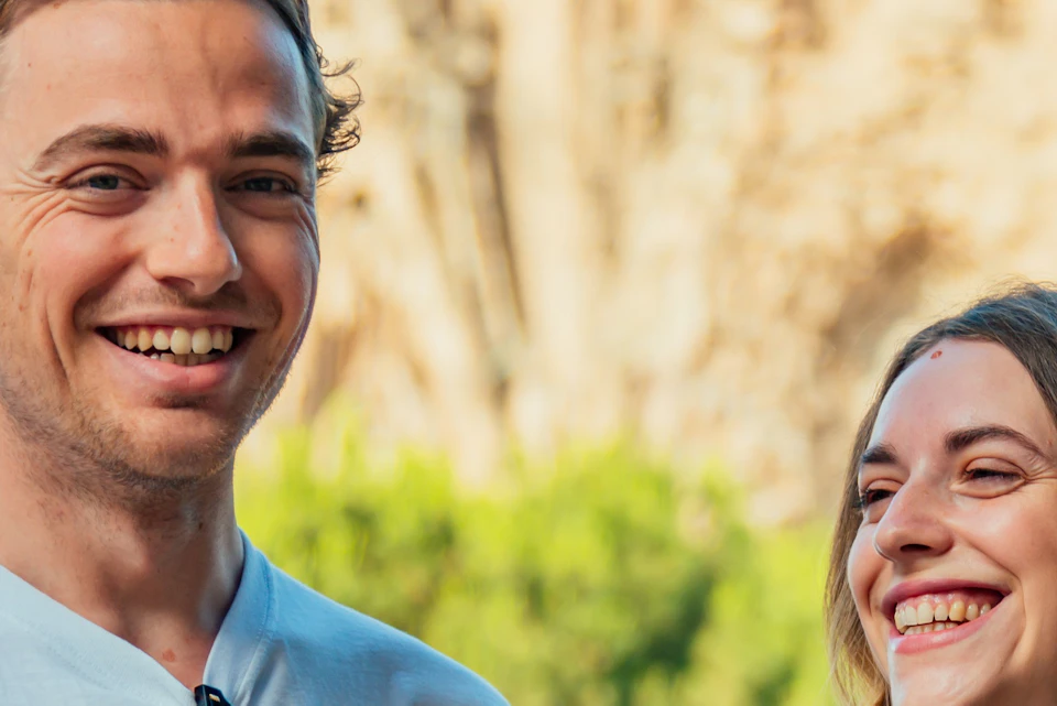 Two smiling people, one wearing a white shirt and sunglasses, the other in a black shirt, standing in front of a blurred background.