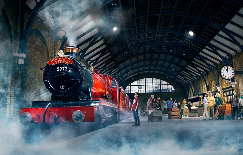 A red steam engine labeled "Hogwarts Express" at a train station with people handling luggage and a clock on the wall.
