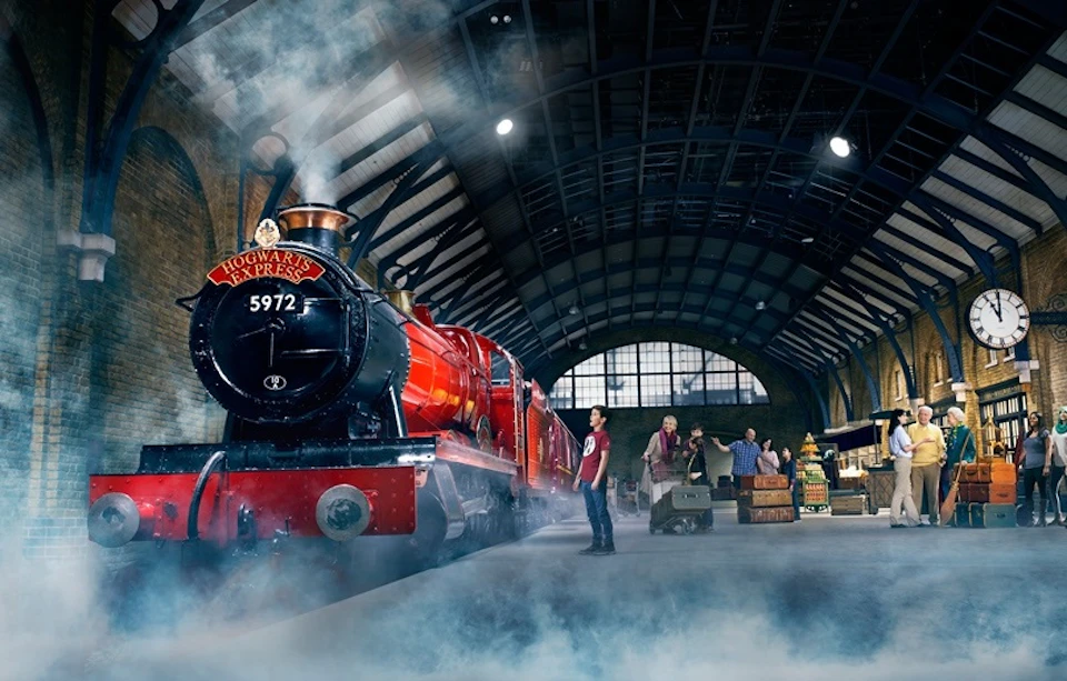 A red steam engine labeled "Hogwarts Express" at a train station with people handling luggage and a clock on the wall.