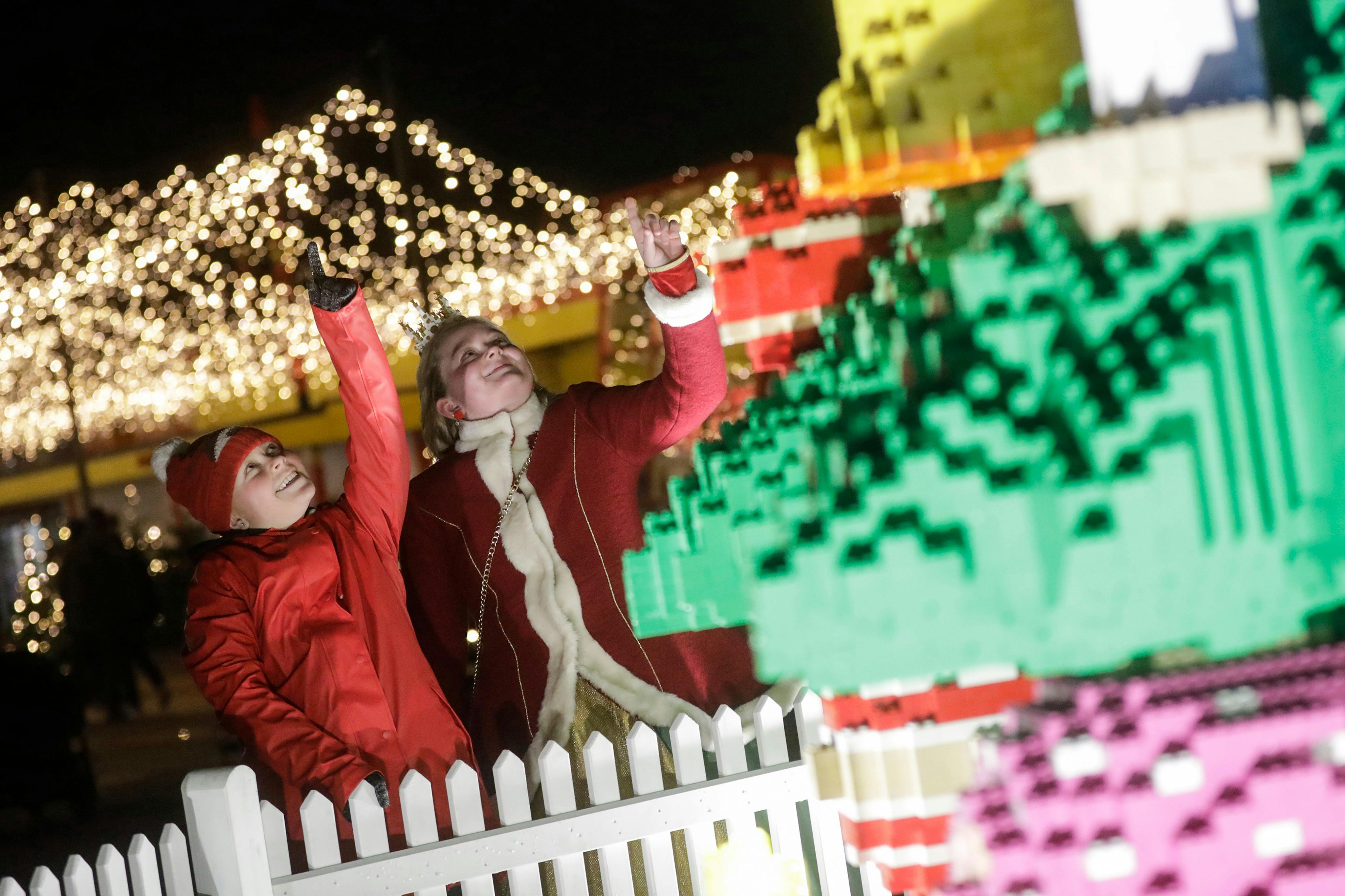 Два человека в праздничной одежде смотрят вверх, стоя рядом с забором с большим дисплеем LEGO и праздничными огнями на заднем плане.