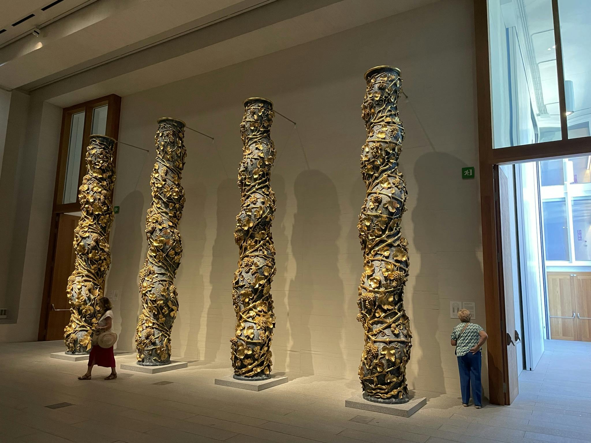 四个由相互交织的材料制成的高大的扭曲雕塑站在一个画廊里，两个人观察着它们。