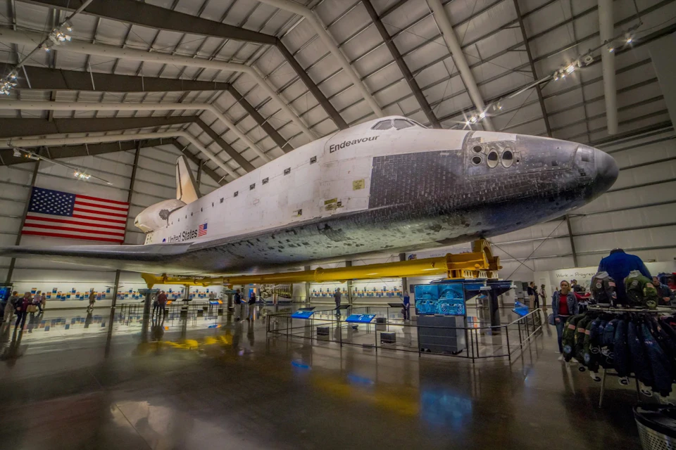 Space Shuttle Endeavour displayed inside a large museum hangar, surrounded by informational exhibits and an American flag on the wall.