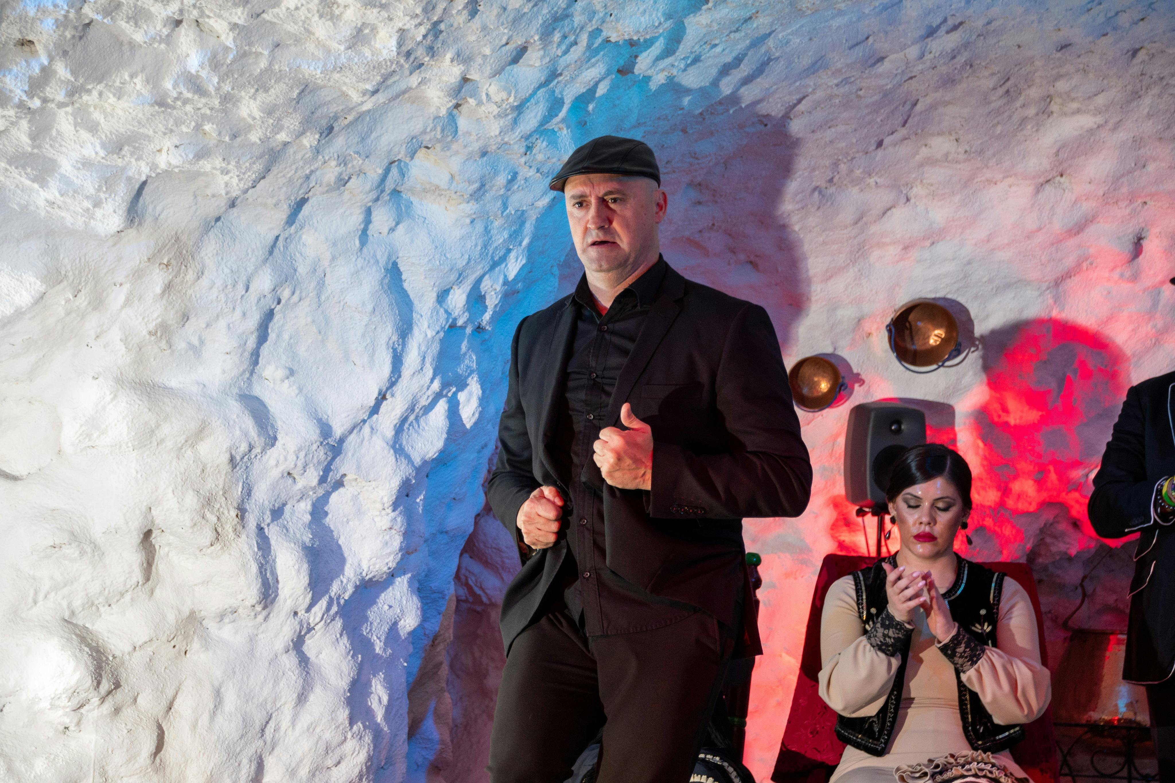 A man in a black outfit and hat stands near a seated woman in a light dress, performing in a cave-like setting with colorful lighting.