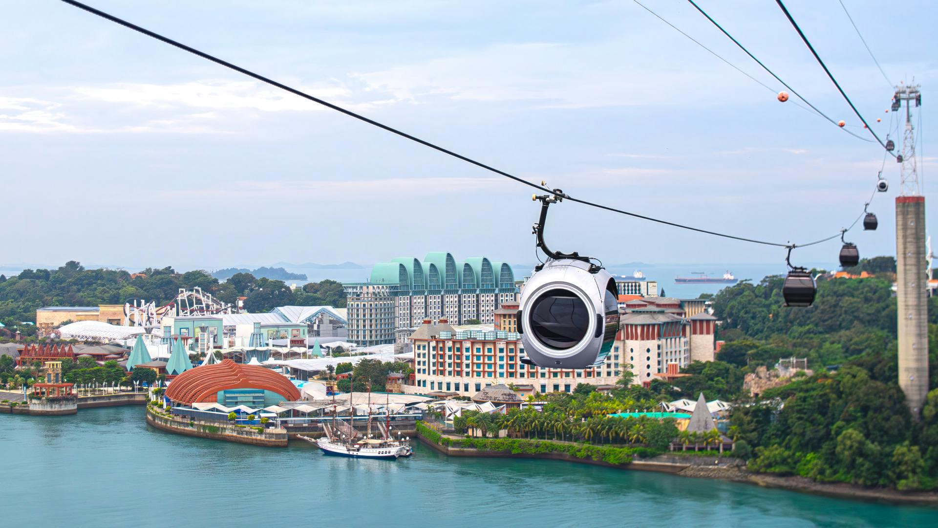 A cable car traveling above a waterfront area with buildings, greenery, and ships in the background.