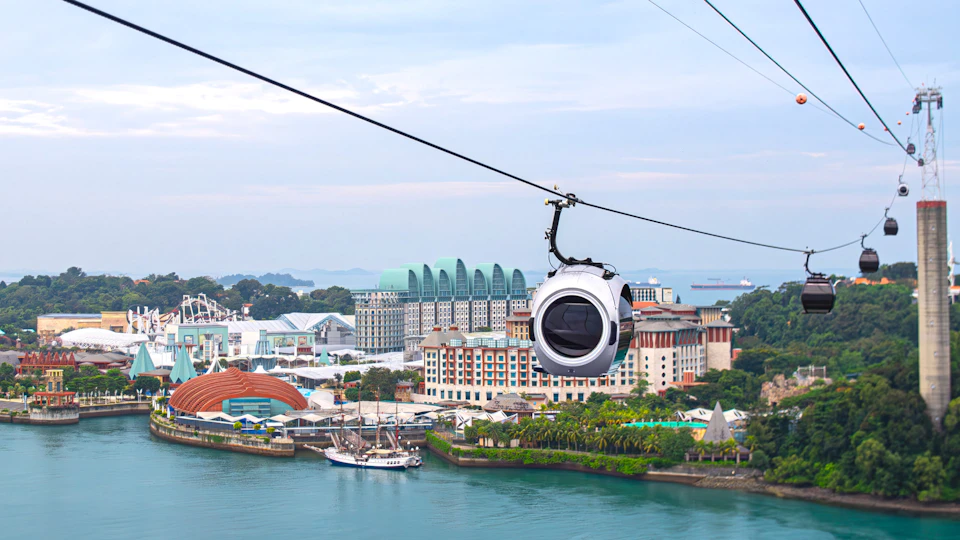 A cable car traveling above a waterfront area with buildings, greenery, and ships in the background.