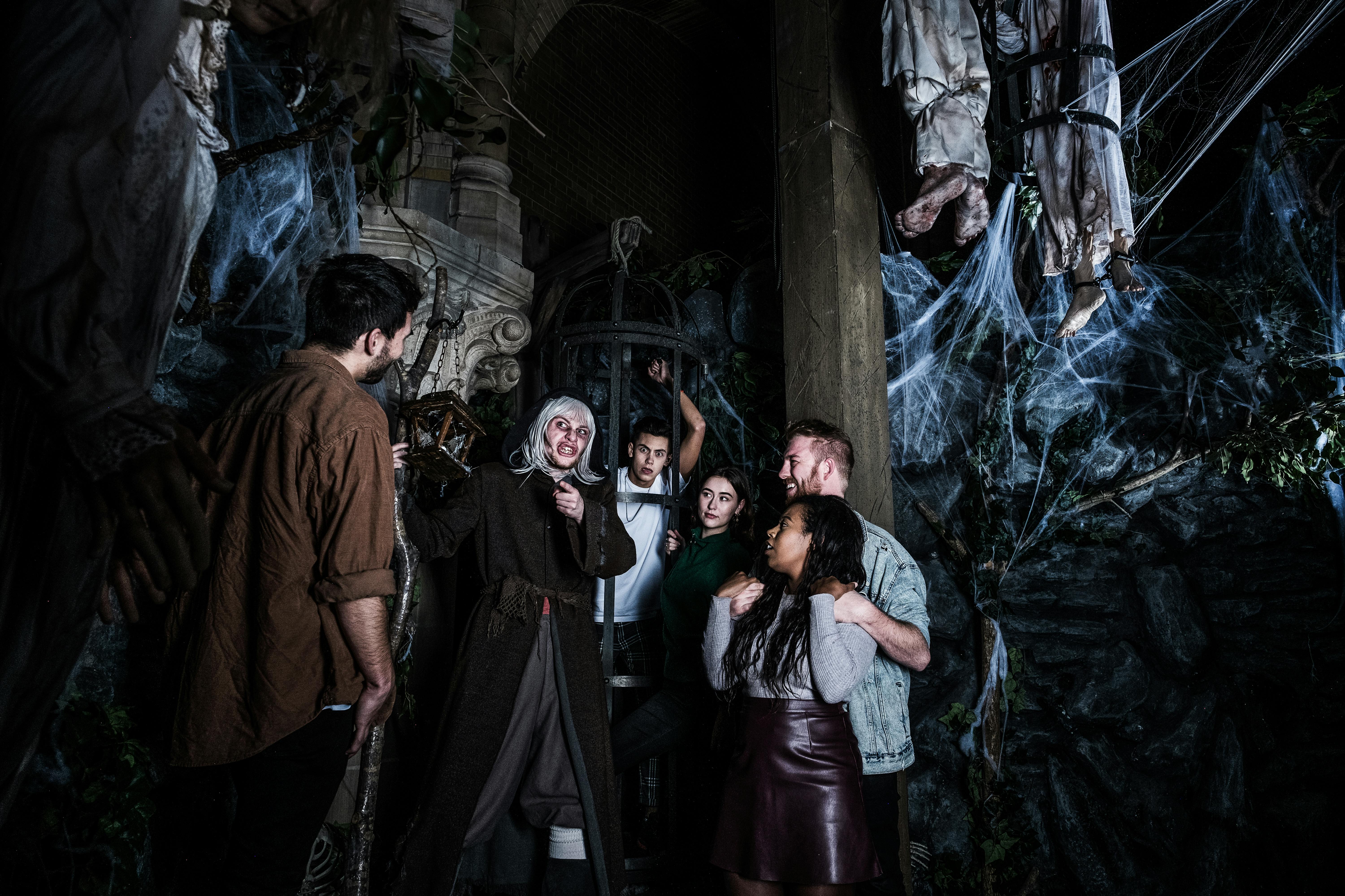 Ludzie stojący wokół upiornego, wypełnionego pajęczyną lochu z osobą w kostiumie trzymającą latarnię.