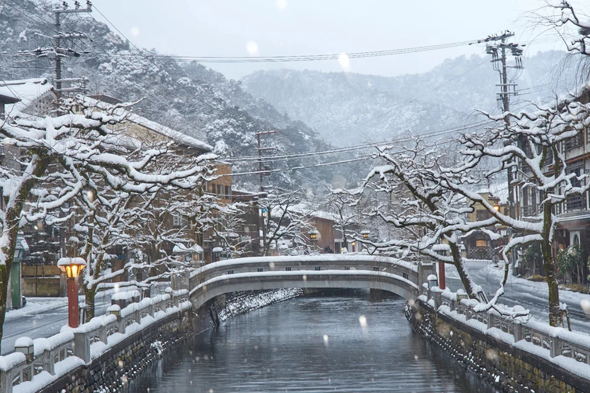 A snow-covered bridge spans over a calm river, surrounded by wintery trees and buildings, with mountains in the background.