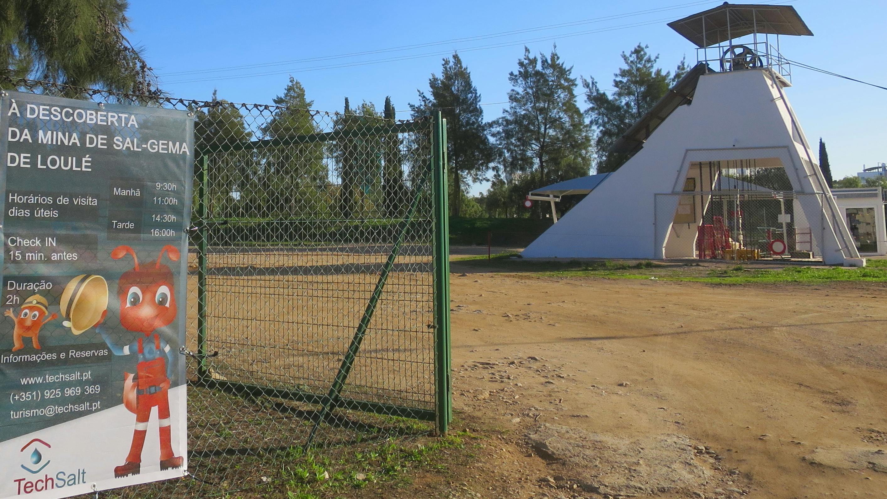 Ανοίξτε την πράσινη συρμάτινη πύλη φράχτη σε έναν χωμάτινο χώρο με δέντρα και ένα λευκό κτίριο στο παρασκήνιο κάτω από έναν καταγάλανο ουρανό.