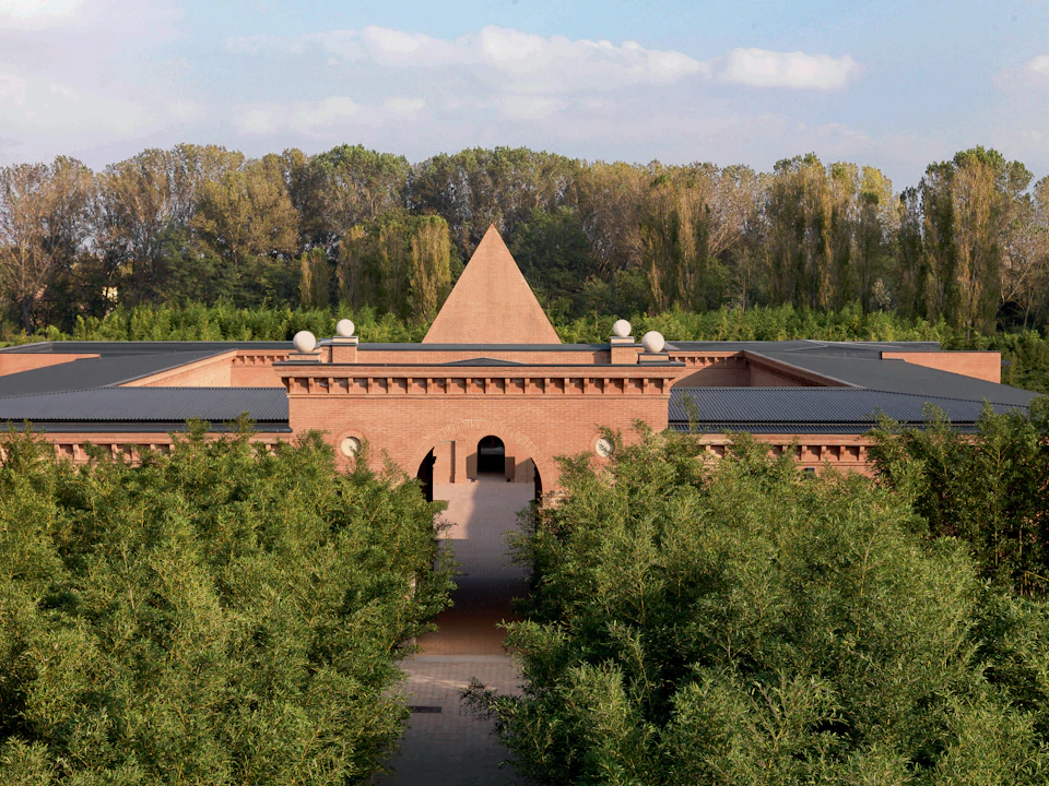 A building with a pyramid-shaped roof and brick facade is set amid lush green trees and dense foliage; a cloudy sky is overhead.