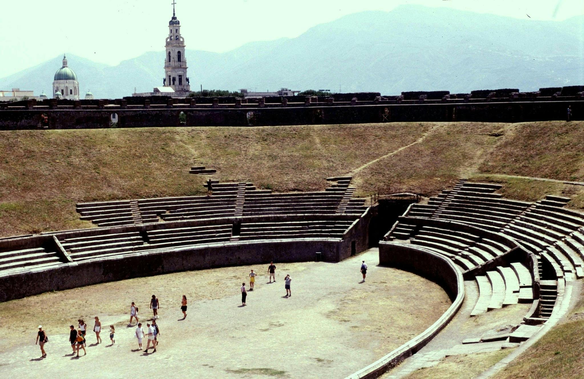 Skupina lidí vstoupí do starobylého amfiteátru s odstupňovanými kamennými sedadly s klenutými a zvonicemi v pozadí.