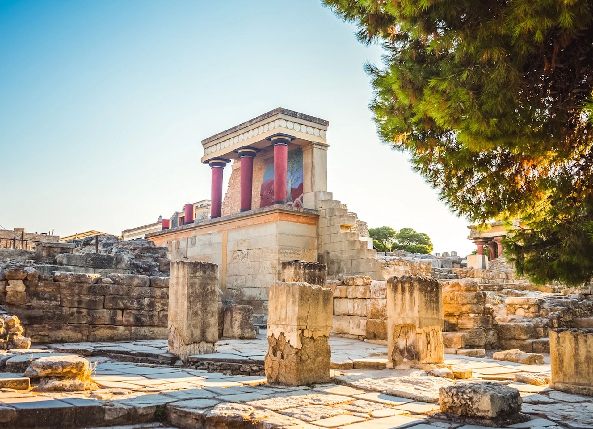 Ancient ruins with columns and a partially restored building featuring red pillars and fresco remnants under a clear blue sky.