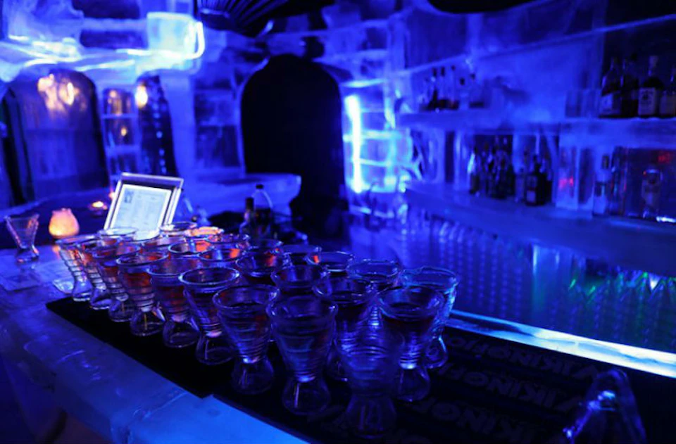 A dimly lit bar with ice walls, numerous glasses on the counter, and illuminated bottles on shelves in the background.